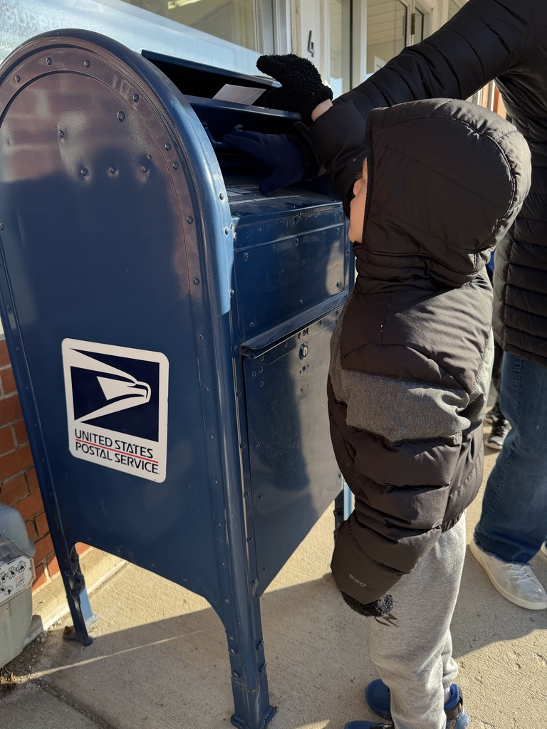 Child wearing a black winter jacket and gloves reaching up to mail a letter in a blue USPS mailbox on a sunny day.