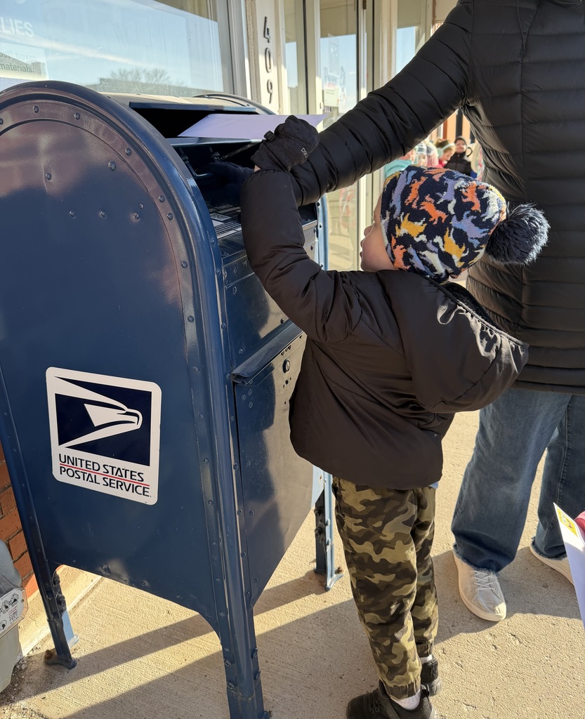 Young boy in a camouflage jacket reaching up to mail a letter in a blue USPS mailbox on a sunny day.