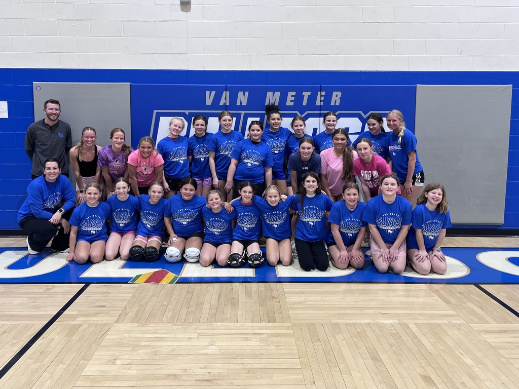 Group of youth softball players in blue “Bulldogs” shirts pose with coaches and older players in a school gym under a “Van Meter” sign.