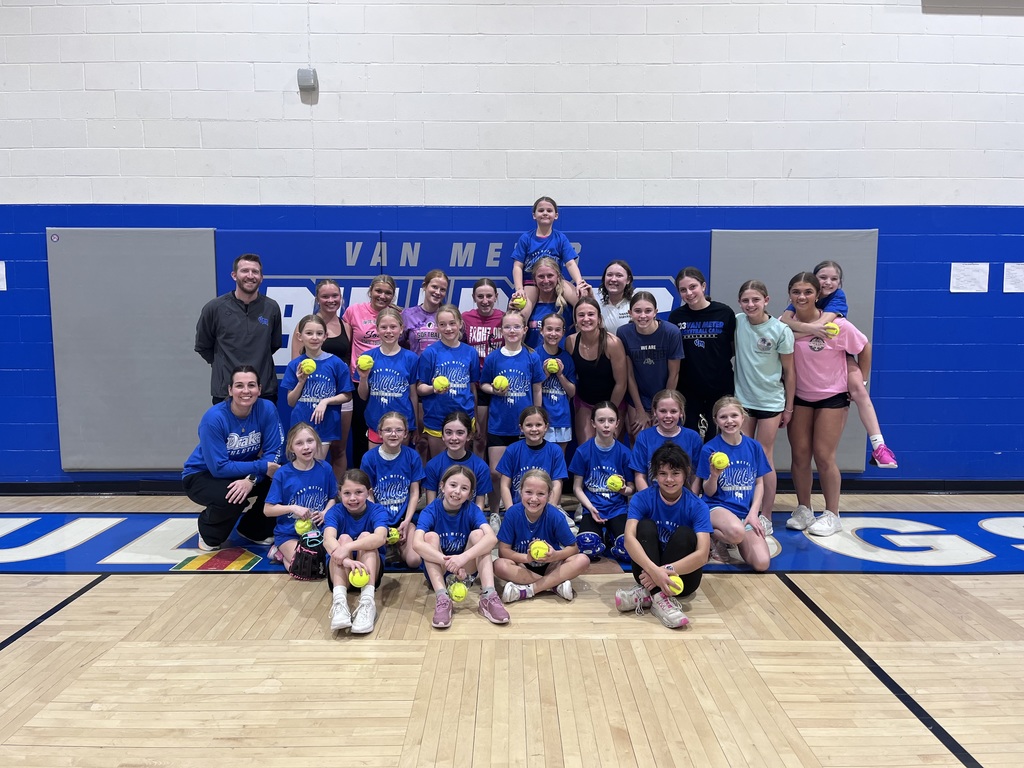 Group of young softball players in blue shirts pose with coaches in a school gym, many holding bright yellow softballs.