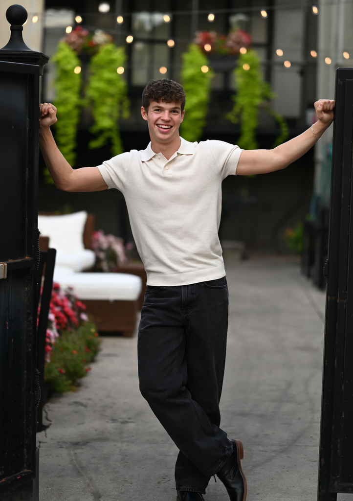 High school student in a white polo shirt and black pants leaning casually against an open black gate in a garden setting with hanging plants and string lights.