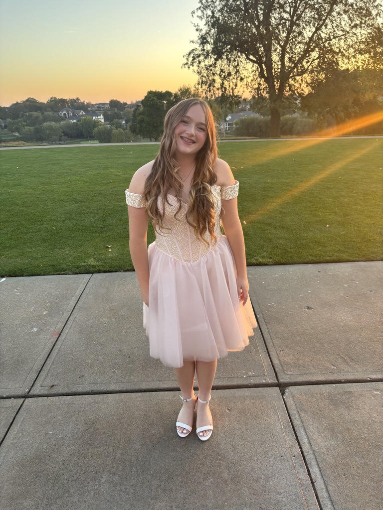 High school student in a light pink off-shoulder dress standing on a sidewalk with a sunset and green lawn in the background.