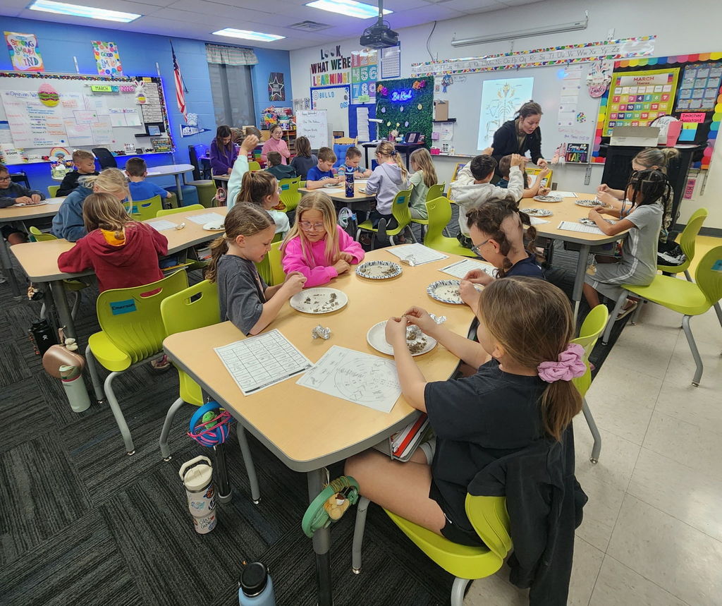 Elementary students sit in small groups, examining materials on paper plates while a teacher circulates in a colorful classroom.