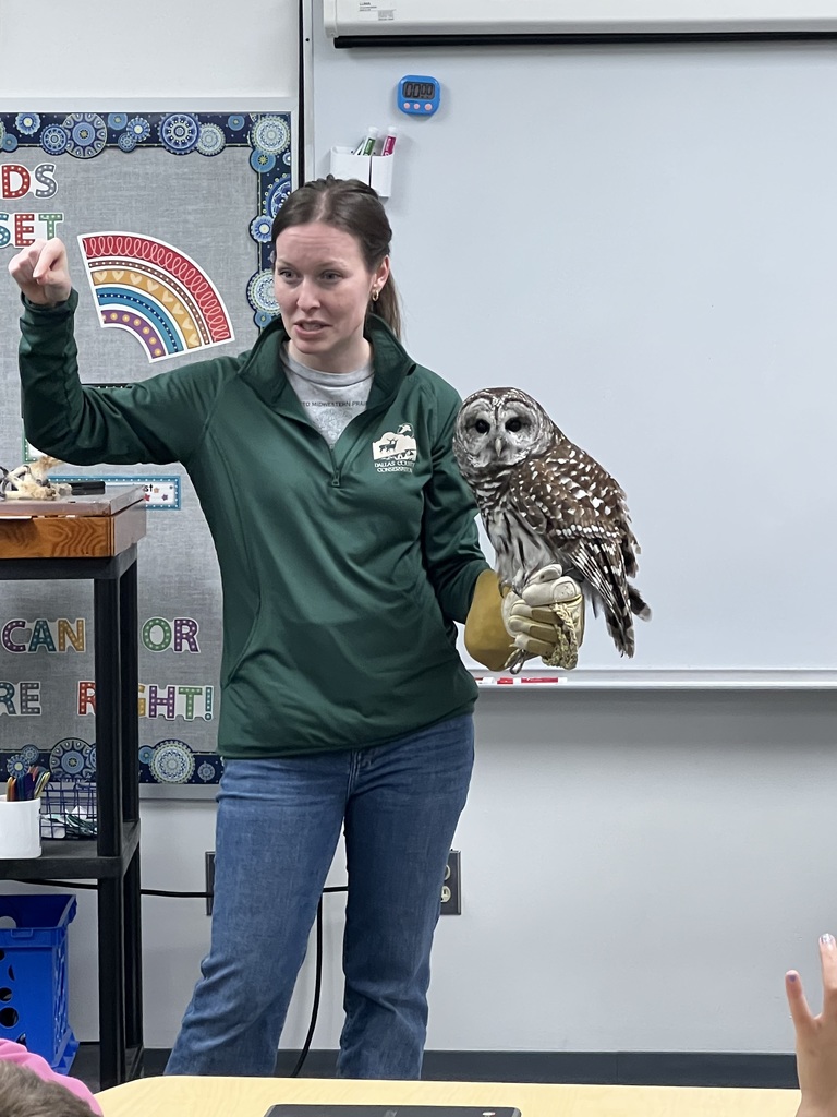 Instructor holds a barred owl on a gloved hand, gesturing while speaking to students at the front of the classroom.
