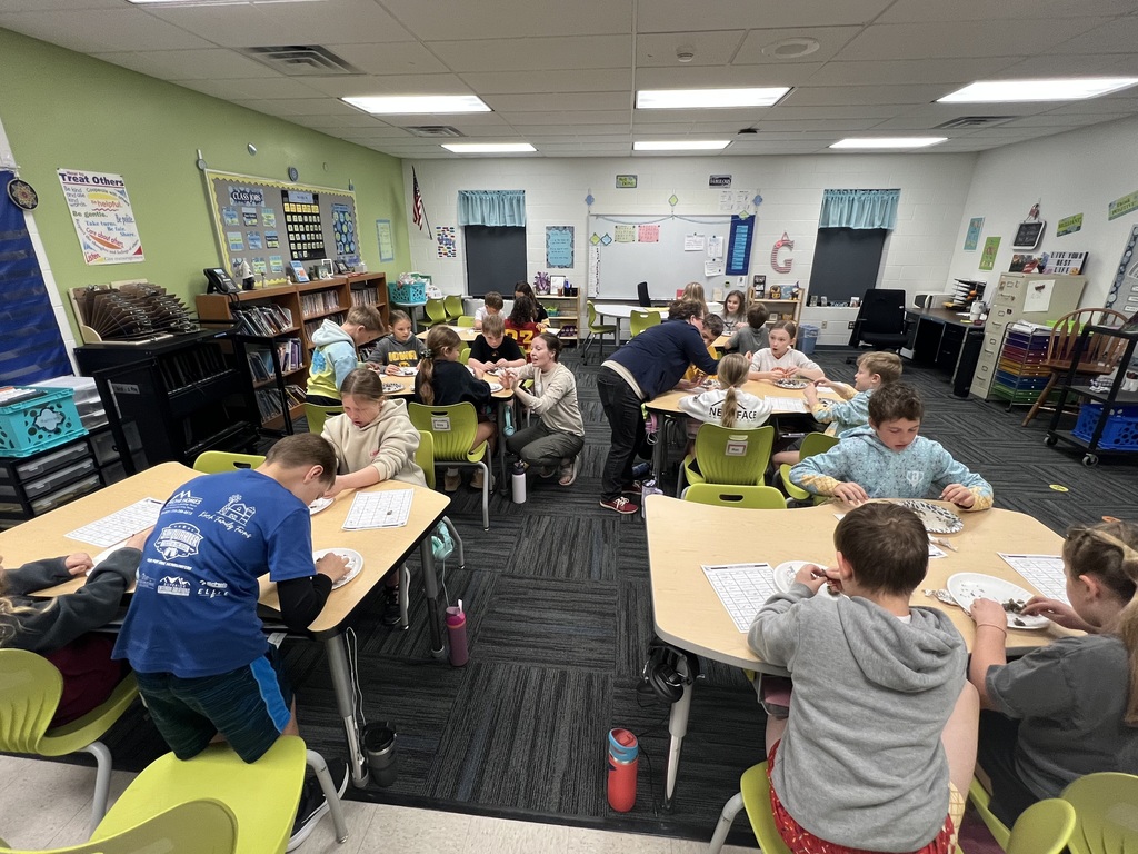 Groups of students collaborate at tables, examining materials on plates as teachers assist around the room.