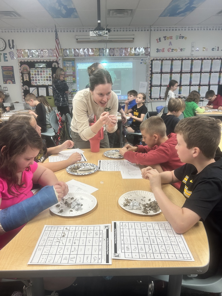 Students work in groups analyzing items on plates while a teacher leans in to guide discussion in a busy classroom.