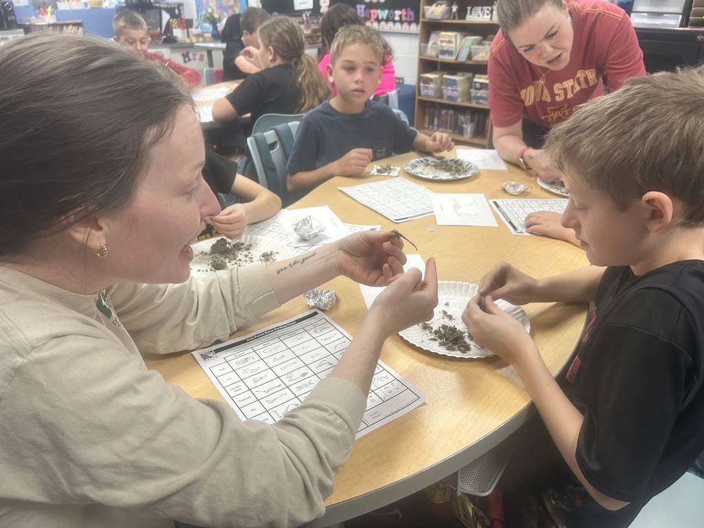 Teacher shows a small specimen to a student at a table as classmates sort and observe materials on paper plates.
