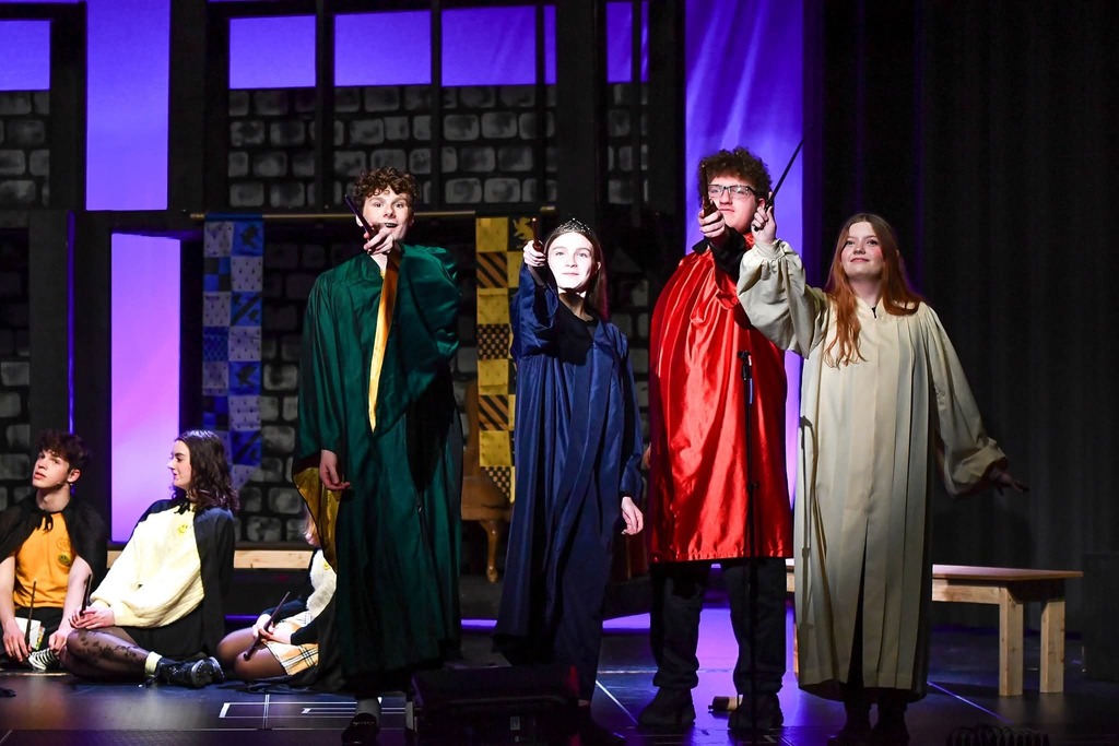 Four students in colored robes stand in a row, pointing wands forward, while others sit behind them onstage.