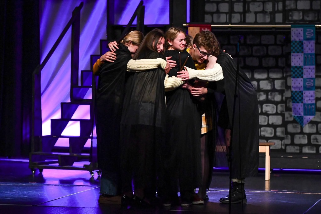 Cast members in dark robes huddle together in an emotional group hug onstage, lit by purple and blue theater lights.