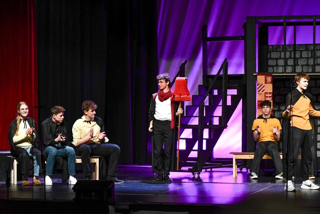Student actor with a red scarf and prop stands center stage while others sit and clap on benches in a school play scene.