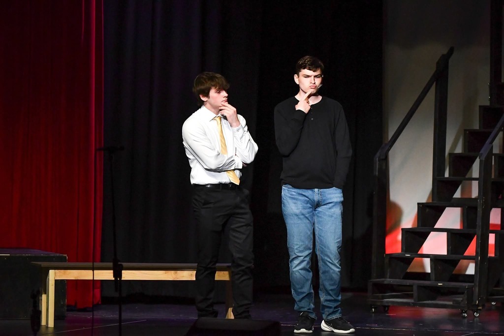 Two students on a dim stage, one in a white shirt and tie, the other in a black shirt, both thinking with hands on chins.