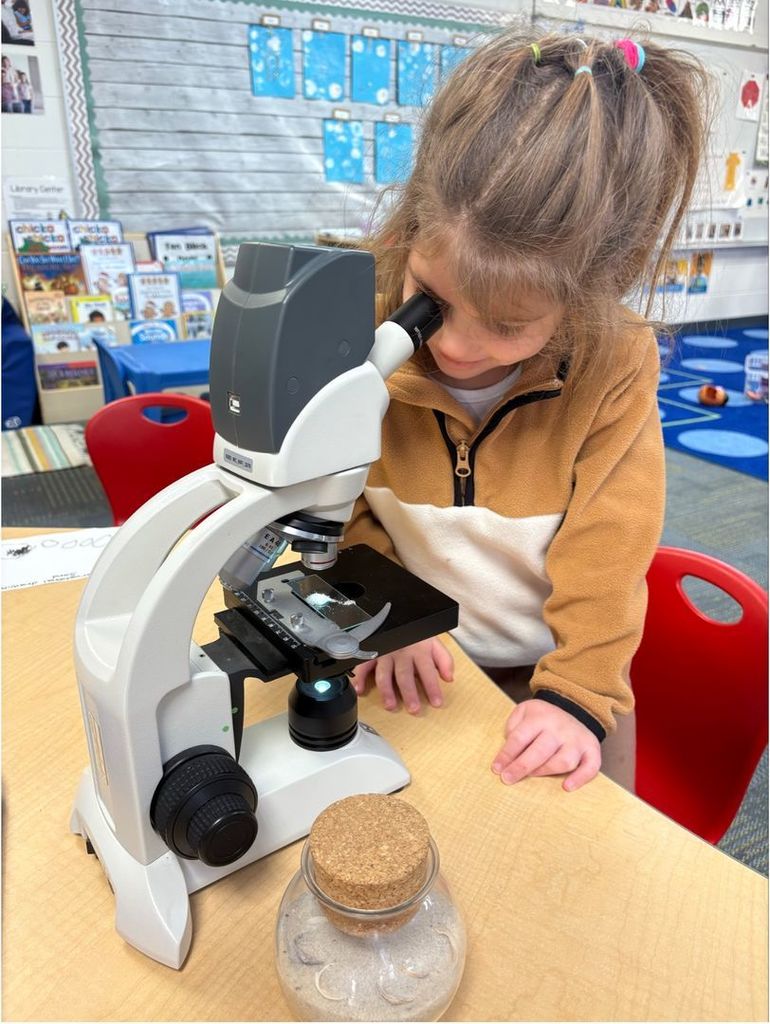 Child looks through a microscope at a table, examining a sample with curiosity in a classroom science activity.