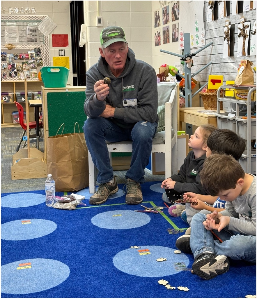 Man in a cap sits on a chair in a classroom, holding a small object while children sit on a blue rug listening attentively.