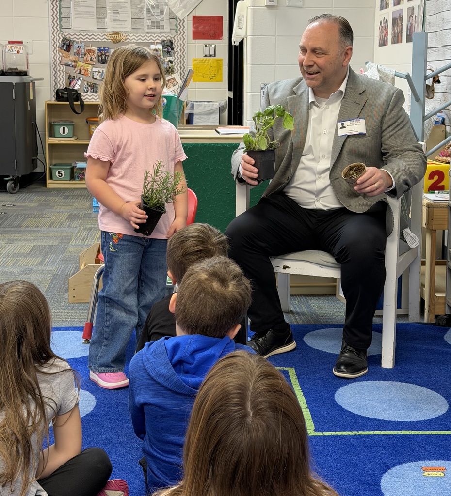 Man in a suit holds a potted plant while a child presents another plant to classmates seated on a classroom rug.