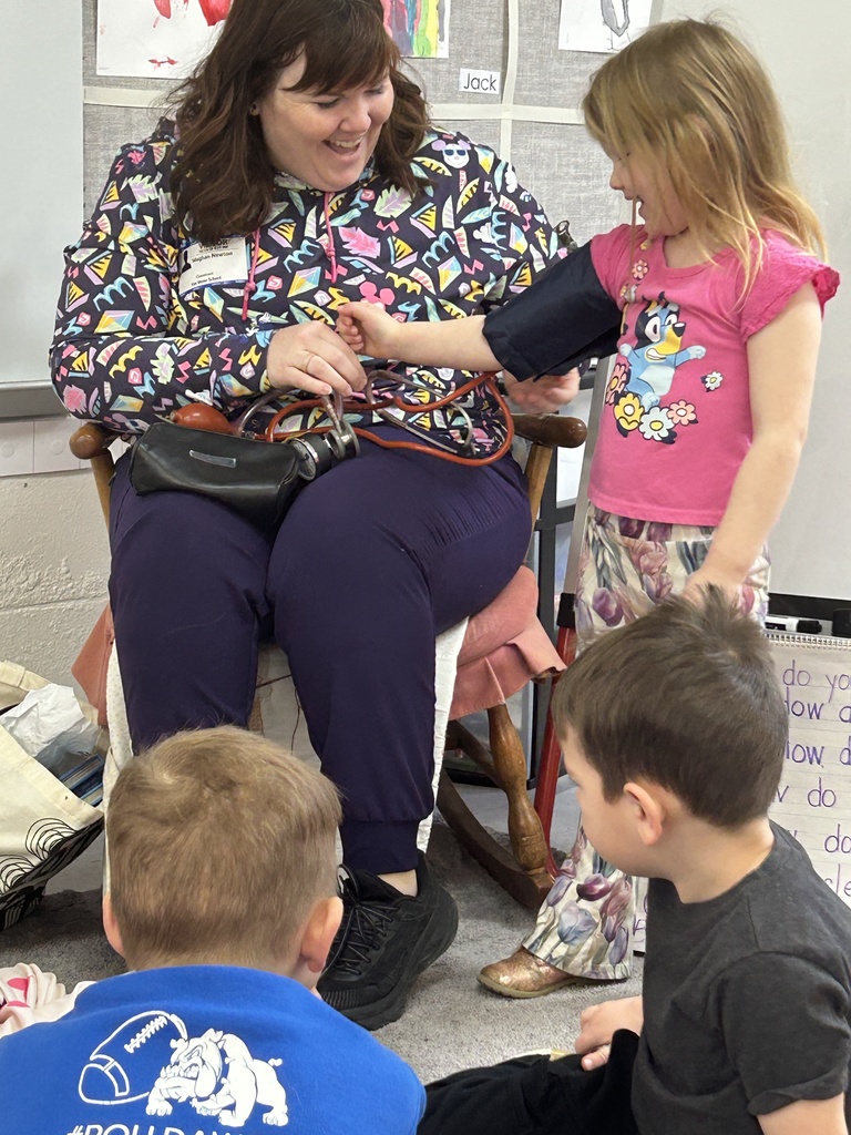 Teacher smiles while a child uses a blood pressure cuff on her arm, as other children watch during a classroom health activity.
