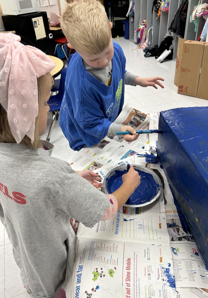 Two children paint a large object blue on a table covered with newspaper, concentrating on their collaborative art project.