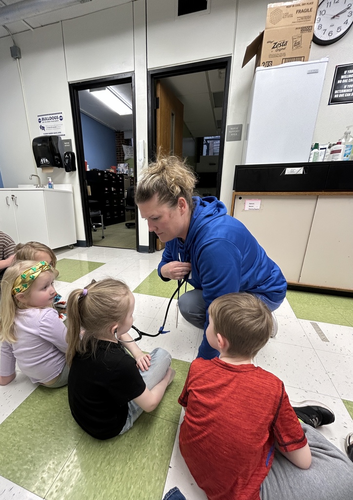 Adult kneels with a stethoscope, guiding children seated on the floor as they listen and learn about how it works.