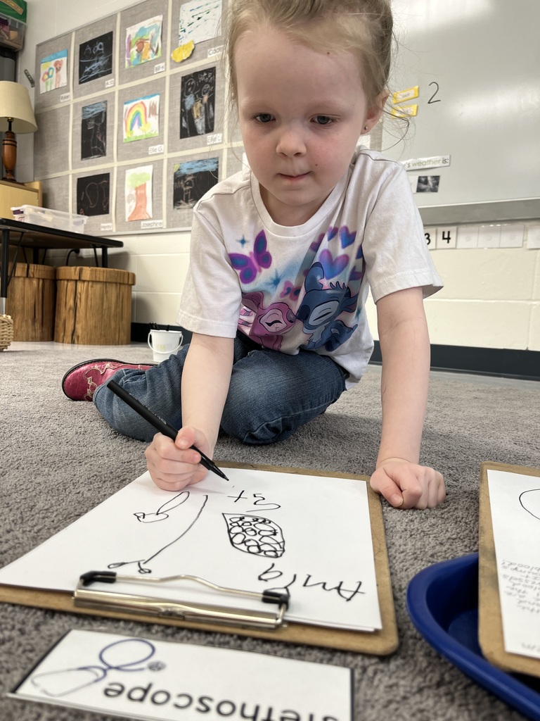 Young child sits on the floor drawing on a clipboard with a marker, focused, with colorful artwork displayed on the wall behind.