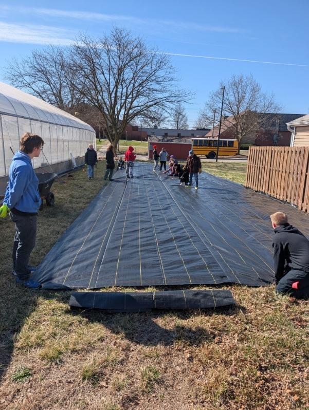 Group of people spreading a large black tarp on the ground near a greenhouse on a sunny day.