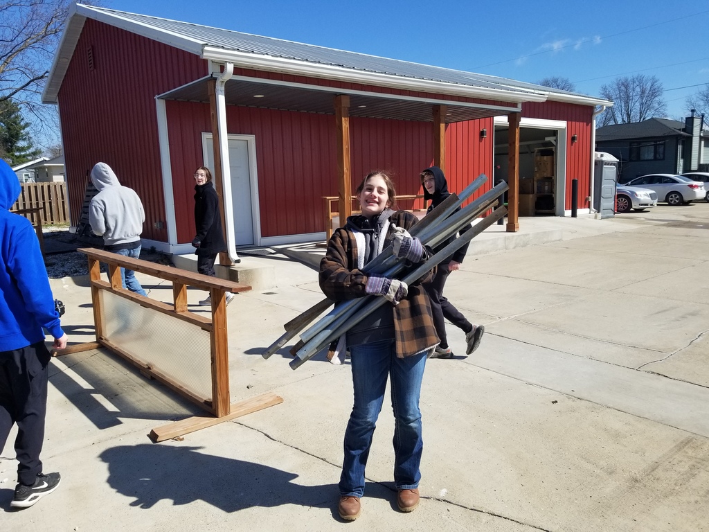 Person carrying metal beams in front of a red barn with others walking nearby on a sunny day.