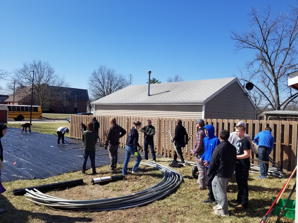 Group of people working together outdoors near a fenced yard with coiled pipes on the ground under a clear blue sky.