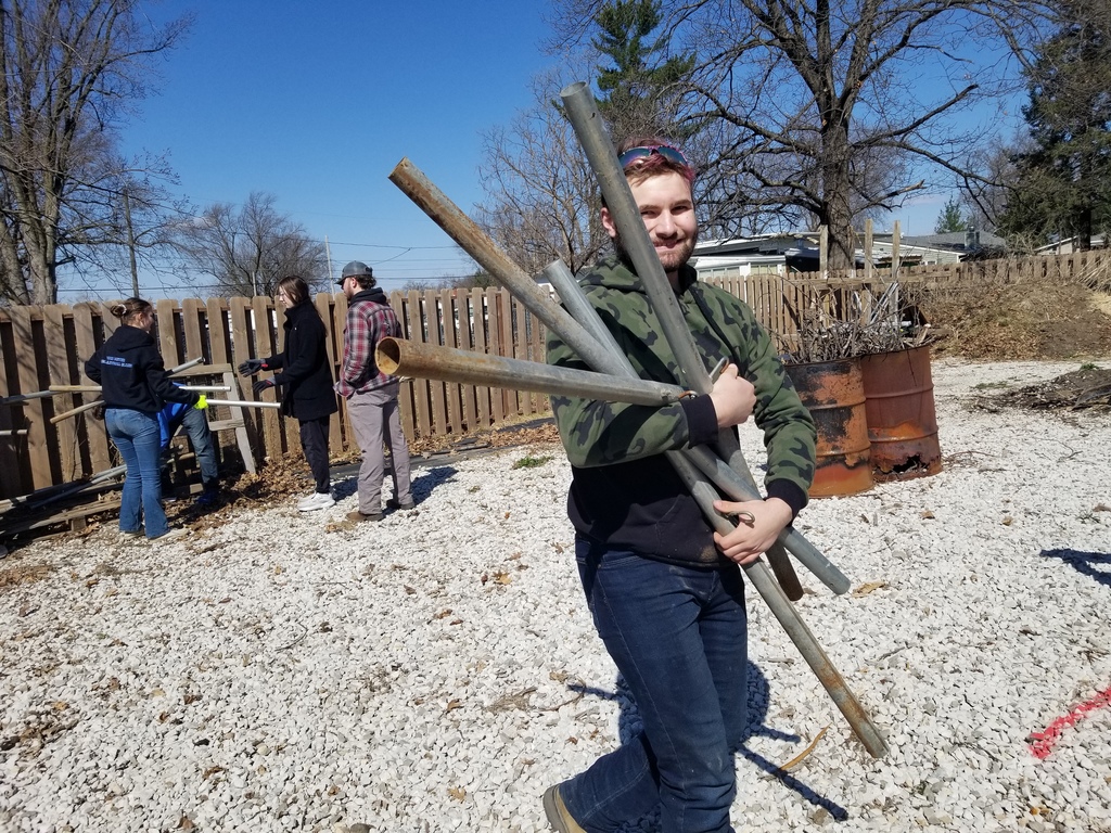 Person carrying several long metal pipes outdoors on a sunny day with others working near a wooden fence.
