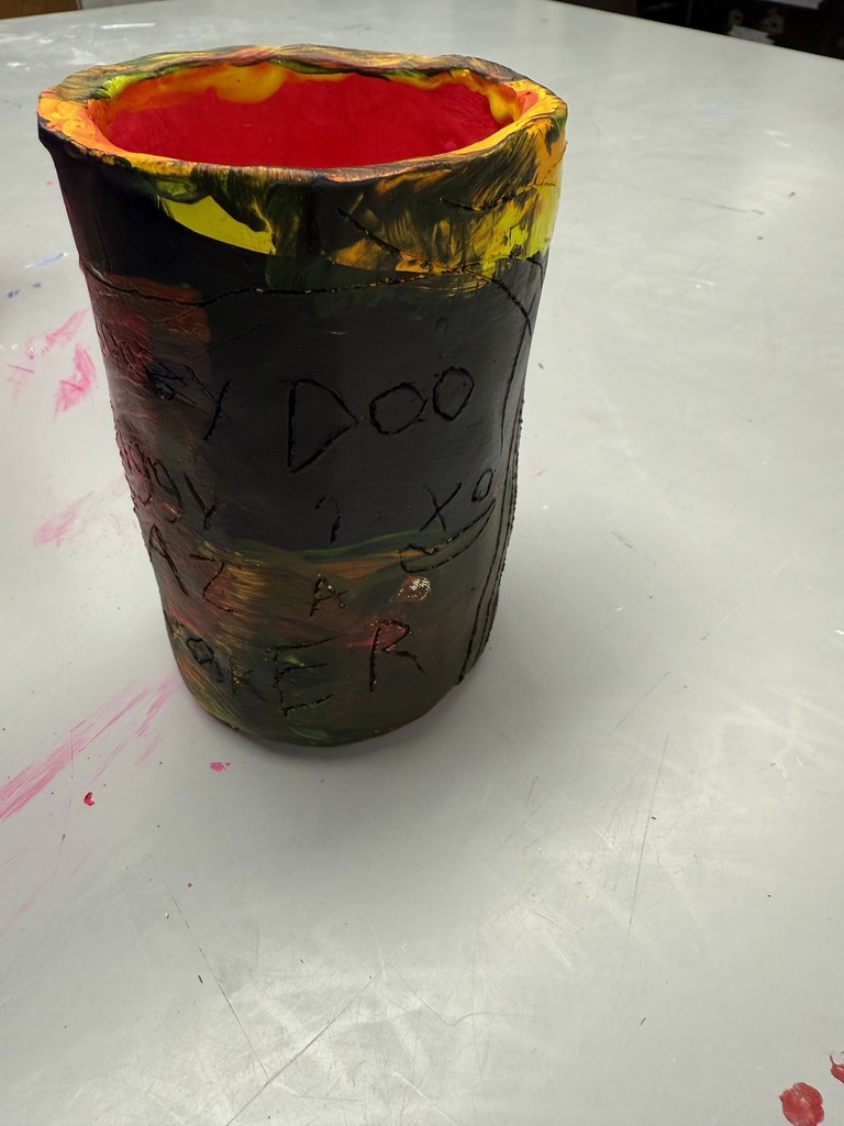 Hand-painted clay cup with carved face and letters, featuring dark exterior and bright red interior on a table.