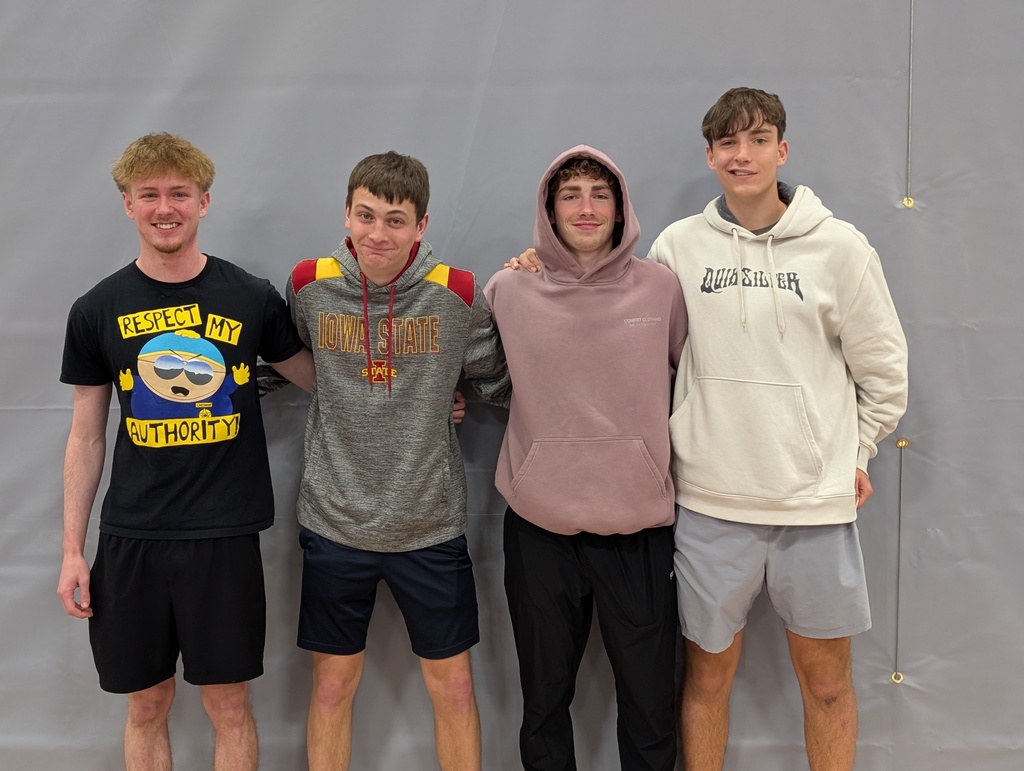 Four teenage boys stand side by side indoors against a gray wall, smiling at the camera with their arms around each other. They are dressed casually in hoodies, T-shirts, and athletic shorts, suggesting a relaxed, school or sports setting.