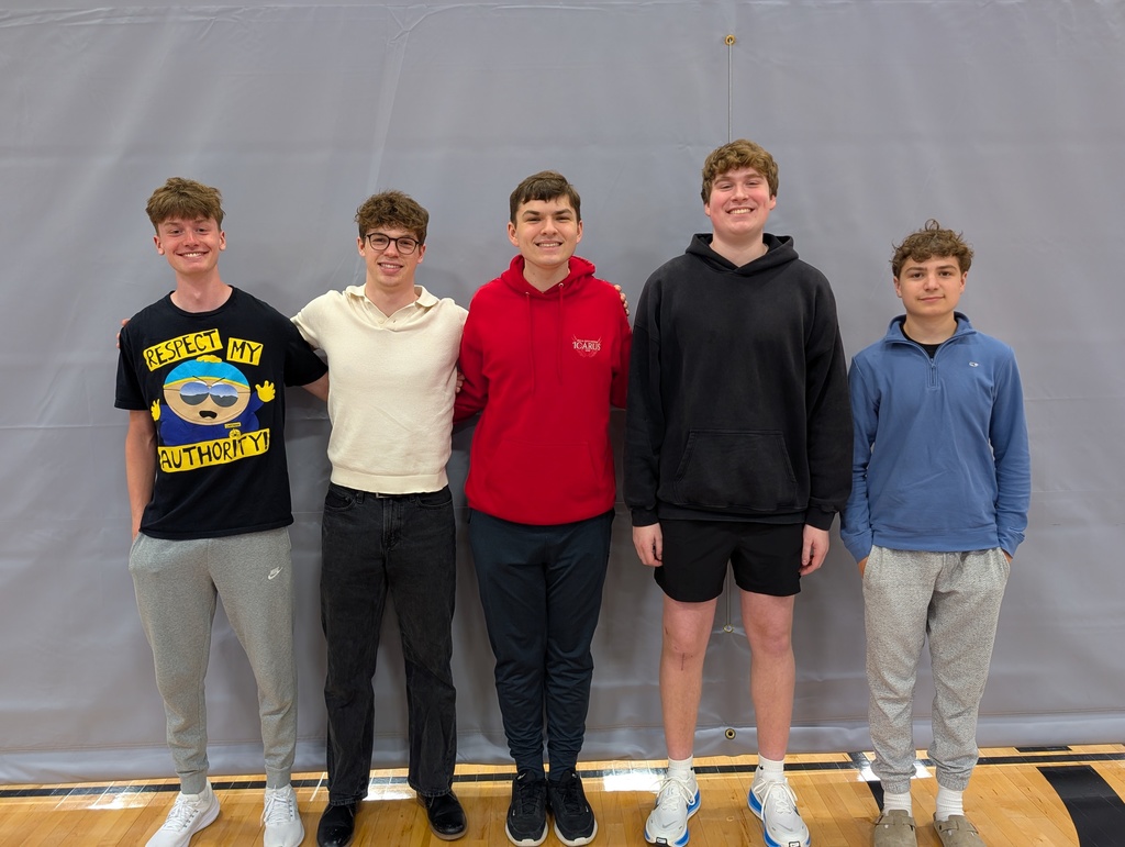 Five teenage boys standing side by side indoors against a gray backdrop on a wooden floor, dressed casually in hoodies, sweatshirts, and sneakers.