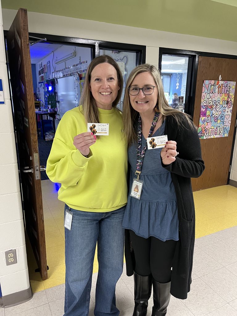 Two women standing in a school hallway holding up identification cards and smiling at the camera.