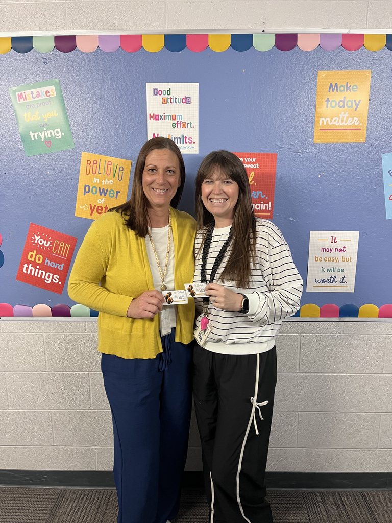 Two women standing in front of a colorful bulletin board with motivational posters, holding an ID card together.