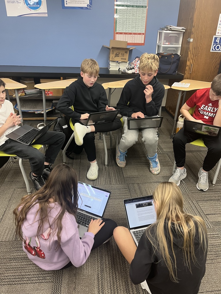 A group of six students working on laptops in a classroom, seated in a circle on chairs and the floor, collaborating on a project.