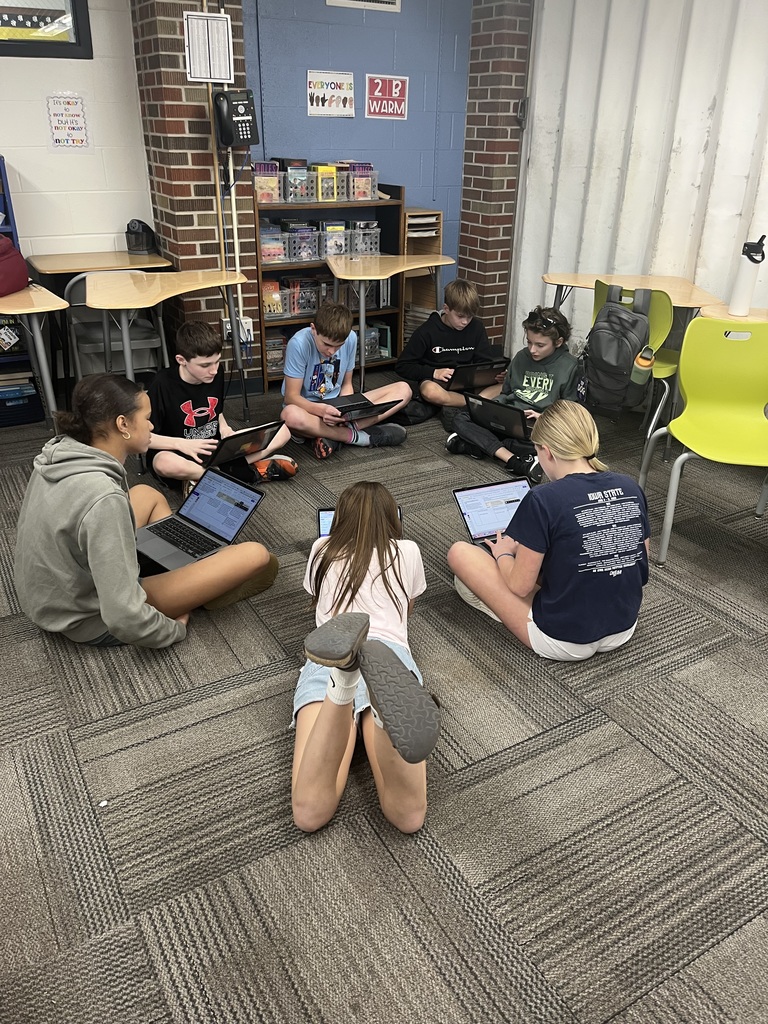 Seven students sitting on a classroom floor working individually on laptops in a circle near desks and shelves.