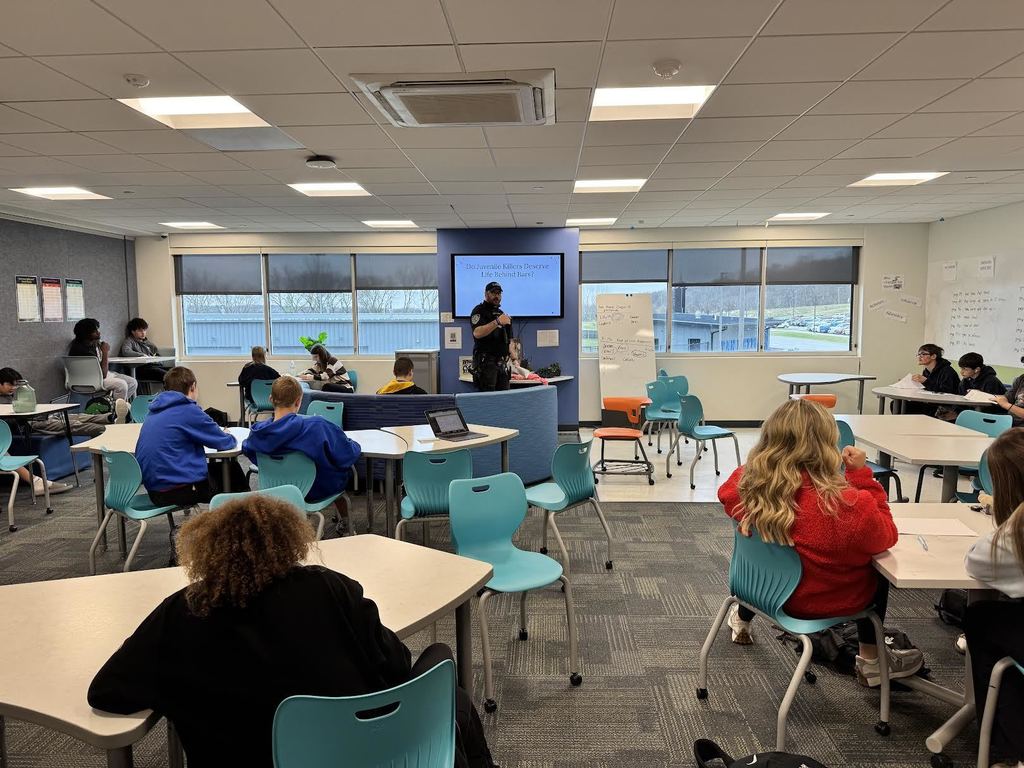 Students seated at round tables in a modern classroom while a teacher presents information on a screen at the front.