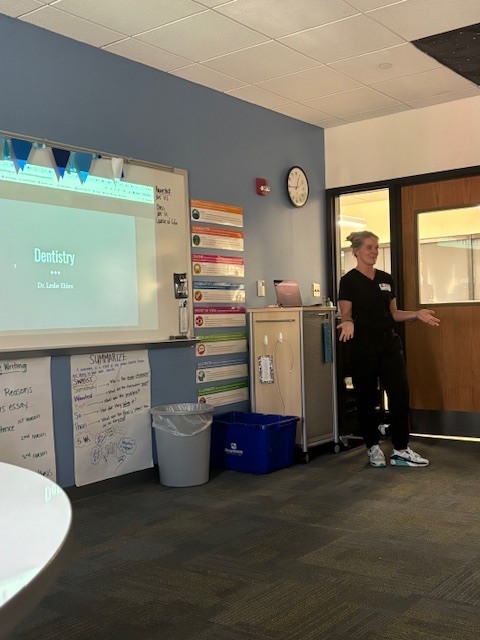 A healthcare professional stands at the front of the classroom presenting about dentistry, with a slide displayed on the whiteboard behind her. Students listen from their seats.