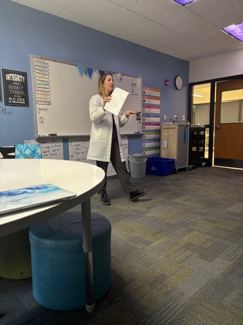 A woman stands at the front of the classroom holding papers and speaking to students, gesturing as she presents. A whiteboard and classroom posters are visible behind her.