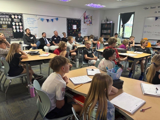 A classroom of elementary students sits at tables, listening attentively to a panel of adult guest speakers seated along the wall. Notebooks, water bottles, and classroom decorations are visible throughout the room.