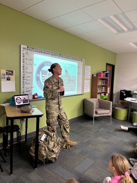 A member of the military in uniform speaks to students seated on the floor, with a backpack and gear placed nearby. A projected image is displayed on the whiteboard behind her.