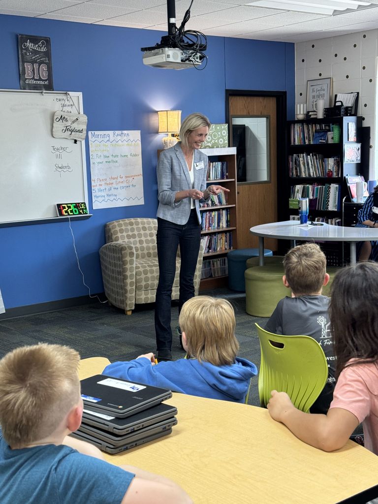 A speaker stands at the front of a classroom, smiling and pointing as she speaks to a group of elementary students at tables. A whiteboard, bookshelves, and classroom posters are visible in the background.