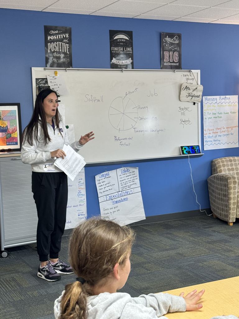 A woman stands near a whiteboard presenting to students, holding papers while explaining a diagram drawn on the board. Students sit at tables facing her in a bright classroom.
