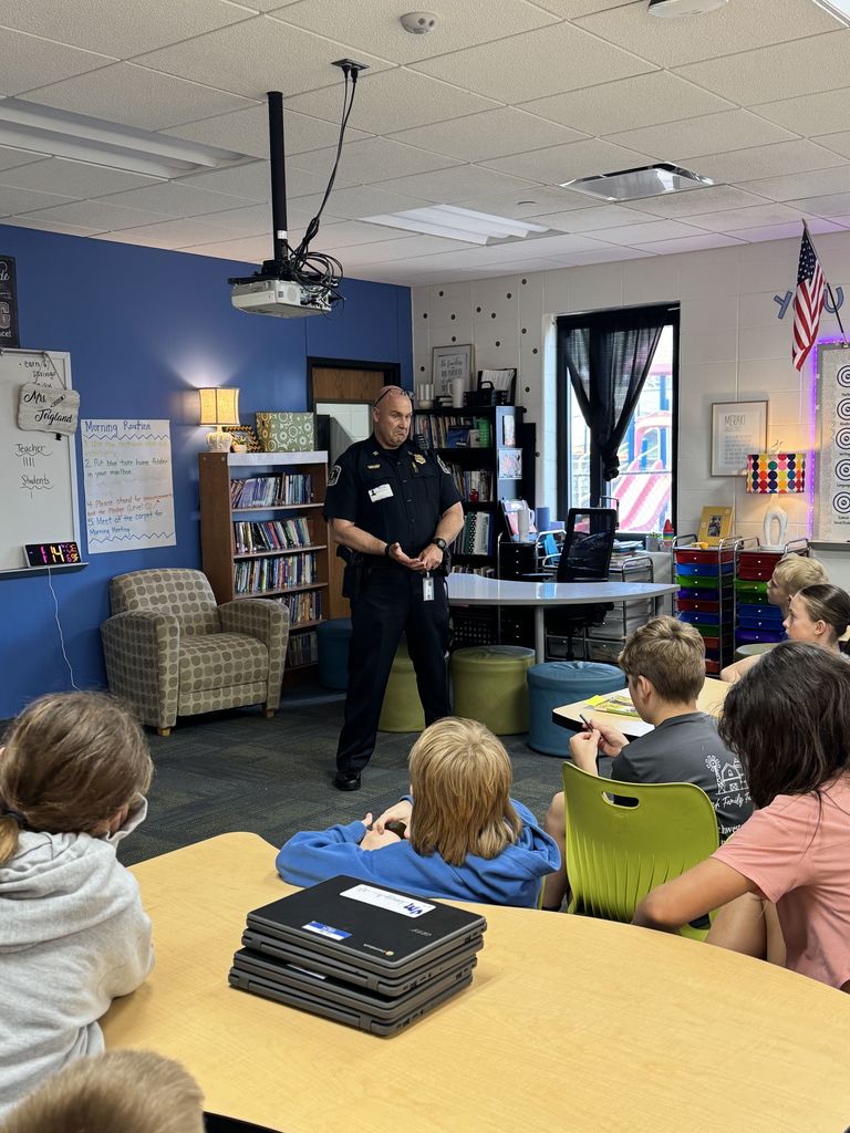 A uniformed police officer speaks to a classroom of students seated at tables, engaging them in a discussion. Bookshelves, classroom posters, and a window are visible behind him.