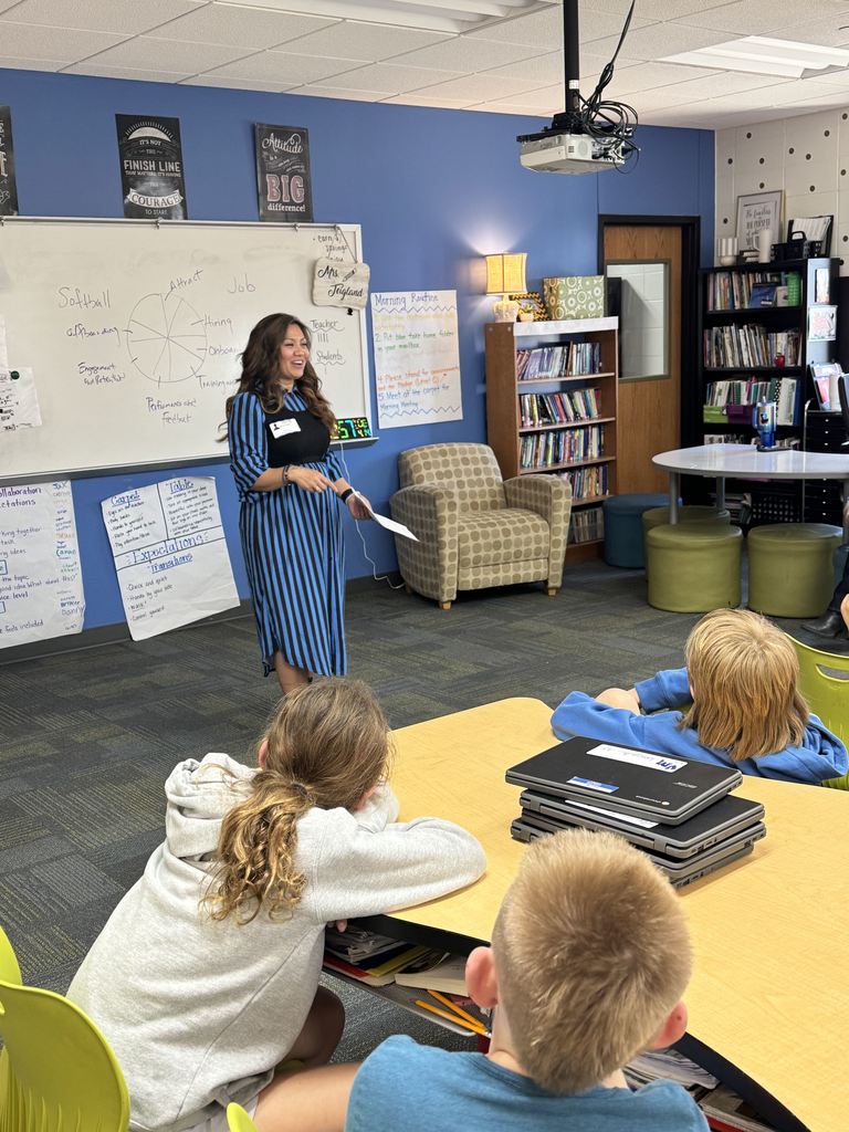 A guest speaker in a striped dress stands at the front of the classroom, smiling and speaking to students seated at tables. The whiteboard behind her displays a diagram and notes.