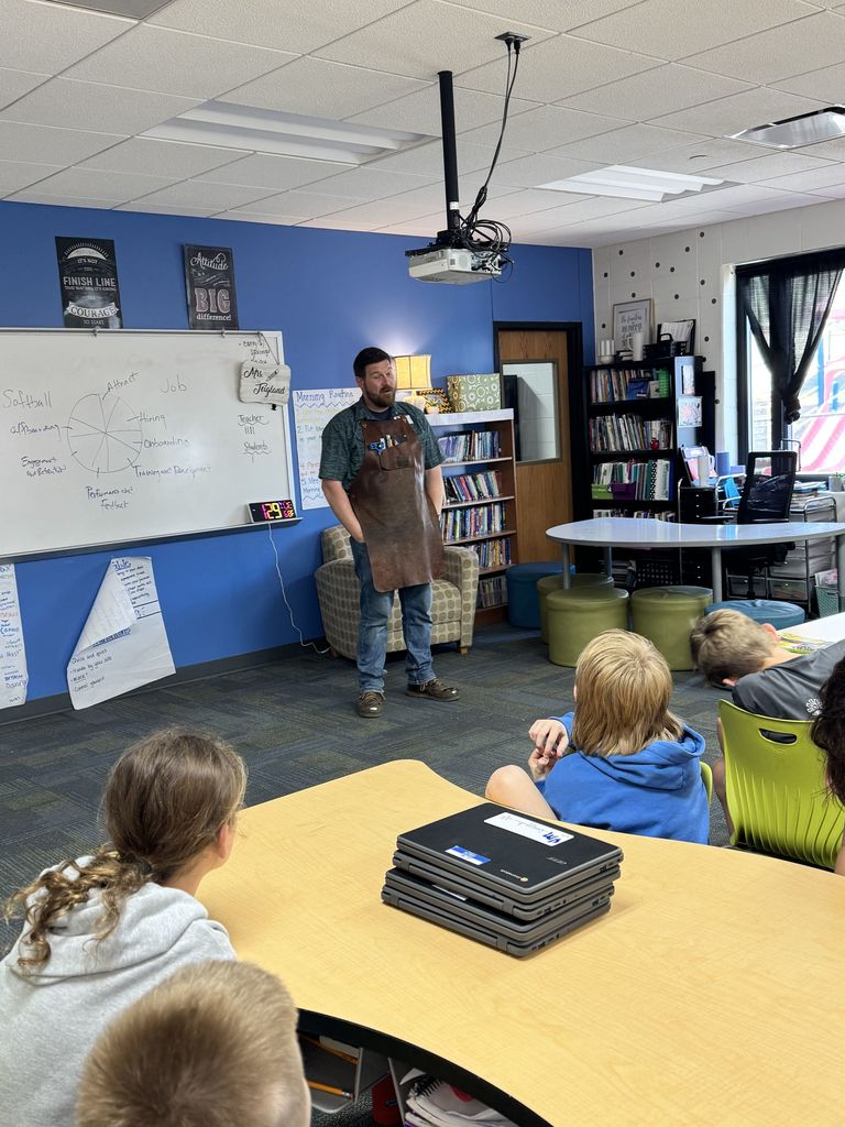A man wearing a work apron stands at the front of the classroom speaking to students, suggesting a hands-on profession. Students sit at tables listening attentively.