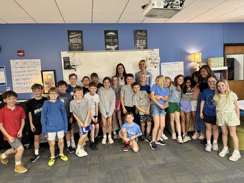A group of elementary students and several adult presenters pose together in a classroom, smiling for a group photo in front of a whiteboard.