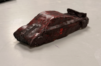 A small, handmade clay car sculpture painted dark red and black, resting on a tabletop.