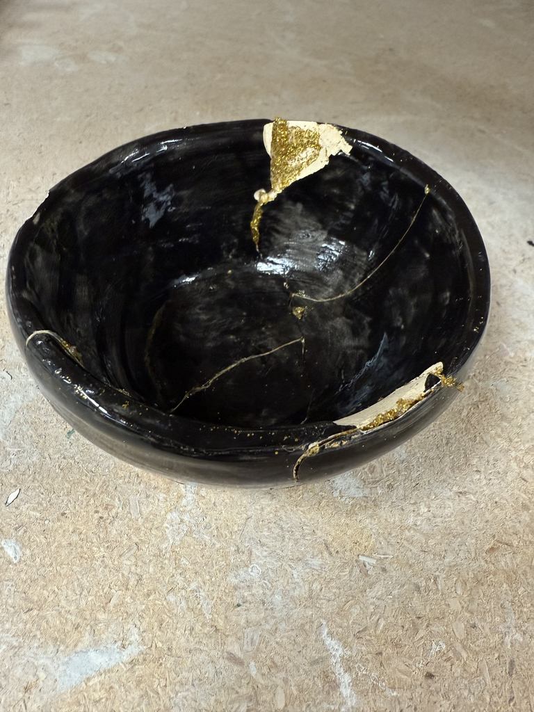 A black ceramic bowl repaired with visible cracks filled with gold-colored material, inspired by a kintsugi style.
