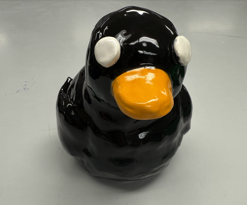 A glossy black ceramic duck sculpture with a bright orange beak and large white eyes, sitting on a neutral surface.