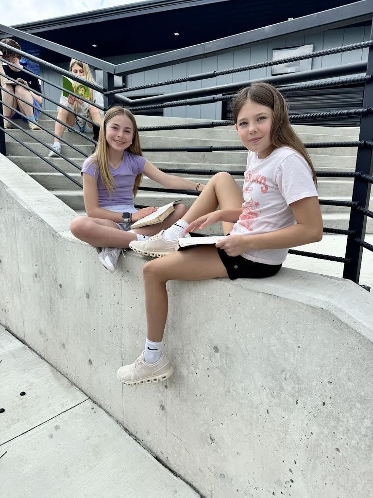 Two girls sitting on a concrete ledge outdoors, each holding a book and engaged in reading near a black metal railing.