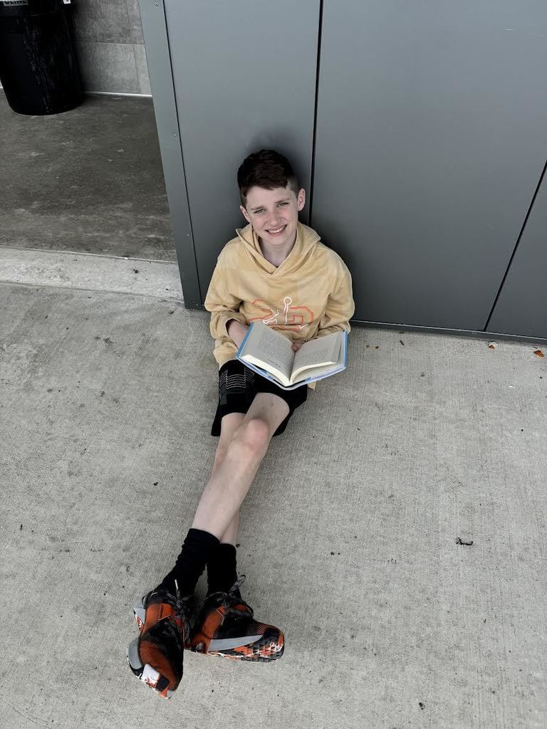 Young boy sitting on concrete floor against a gray wall, reading a book with focused attention.