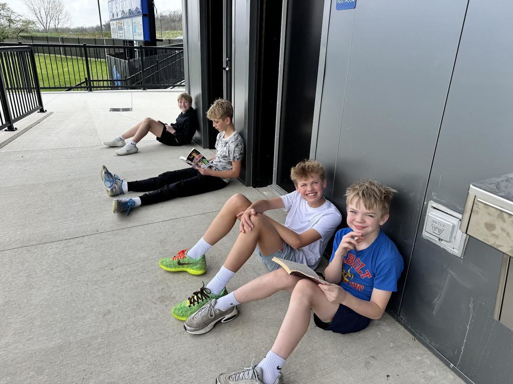 Four boys sitting on a concrete floor outside a building, each engaged in reading a book on a cloudy day.